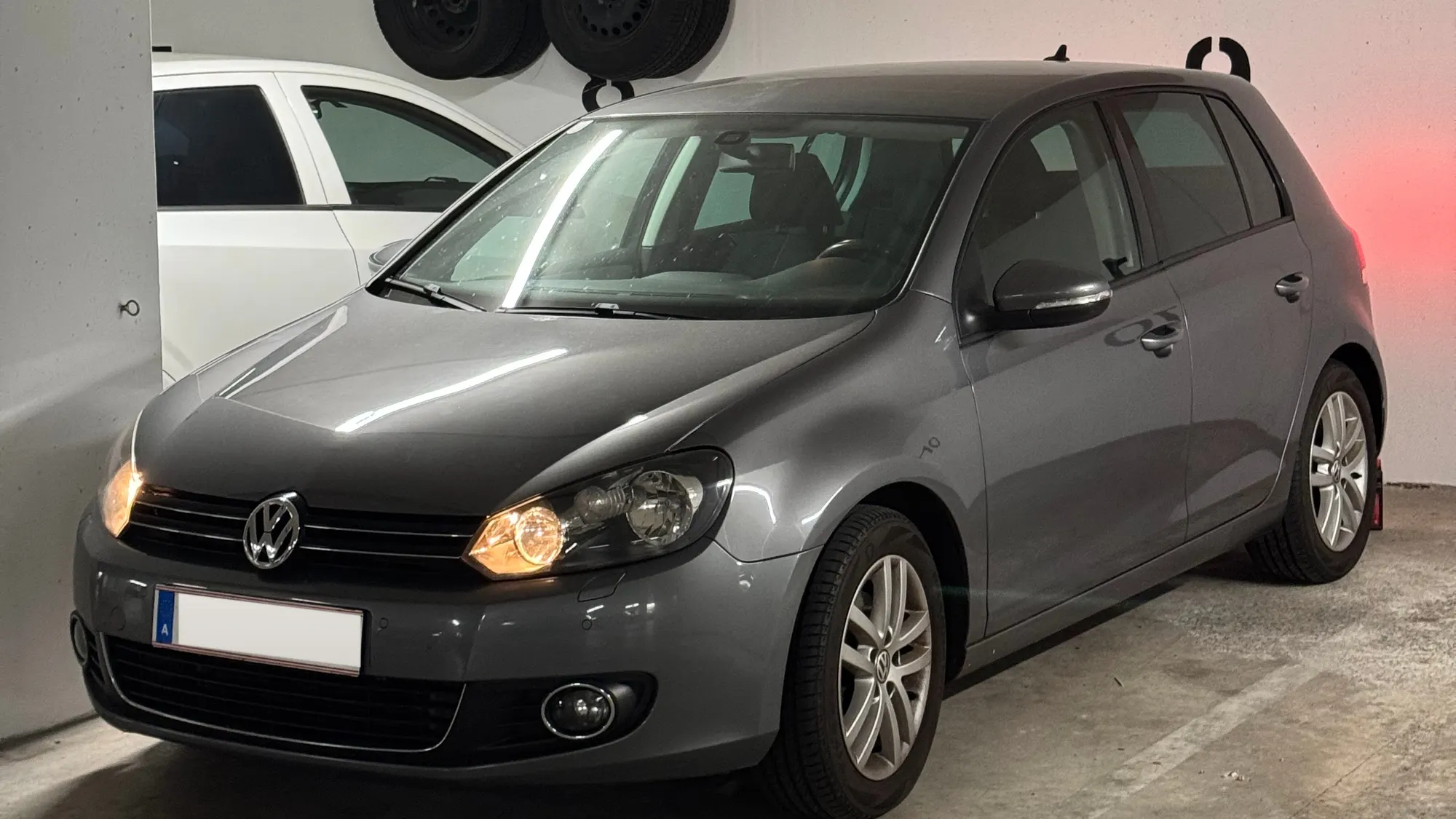 Ein grauer VW Golf mit eingeschaltetem Licht steht in einer Halle, an der Wand hängen Reifen